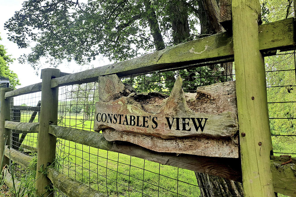A wooden house name sign on a fence says "Constable's View". There is a green field and tree through the fence.